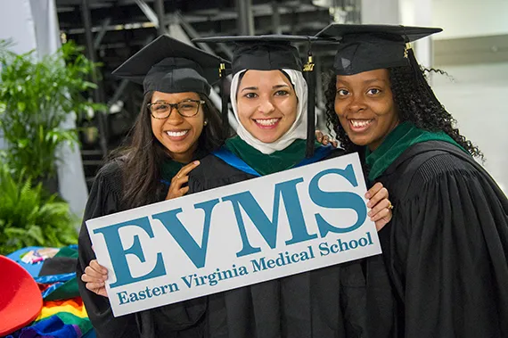 Three MD students wearing graduation robes pose for a picture with a sign depicting the EVMS logo.